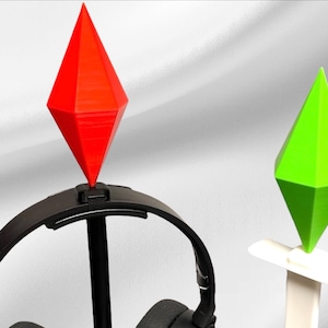 Puede incluir: Un soporte para auriculares negro con una parte superior en forma de diamante rojo y verde. El soporte está diseñado para sostener los auriculares.