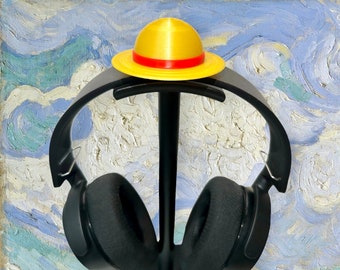 Puede incluir: Un soporte para auriculares negro con dos pares de auriculares negros. El soporte tiene un sombrero amarillo y rojo en la parte superior.