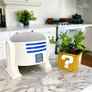 Op de afbeelding: Een witte en blauwe Google Home-luidspreker met R2-D2-thema met een grijze stoffen bovenkant. De luidspreker staat op een wit aanrecht naast een gele vraagtekenblok-plantenpot met groene planten.