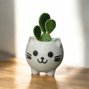 May include: White ceramic cat planter with black eyes and whiskers, holding a small green cactus plant.