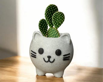 Puede incluir: Maceta de cerámica blanca con forma de gato con ojos y bigotes negros, que contiene una pequeña planta de cactus verde.