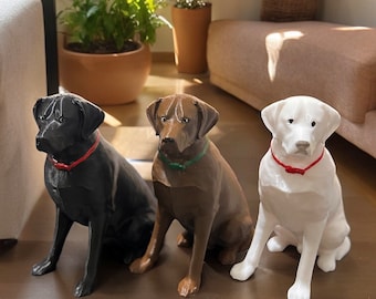 May include: Three Labrador Retriever dog figurines, one black, one brown, and one white, are sitting on a light brown surface. Each dog is wearing a red collar with a bow tie.