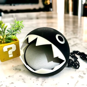 May include: A black and white 3D printed  speaker cover shaped like a character from the video game Super Mario Bros. The cover has a large open mouth with sharp teeth and a single eye. A black chain is attached to the cover.