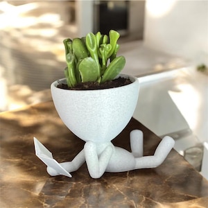 Op de afbeelding: Een witte keramische plantenpot in de vorm van een liggend persoon die een boek leest. De pot heeft een rond hoofd en een kleine plant die eruit groeit.