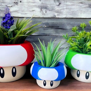 Puede incluir: Tres macetas con forma de hongo en rojo, azul y verde con detalles blancos. Cada maceta tiene una cara de dibujos animados con ojos negros y una pequeña boca. Las macetas están llenas de diferentes tipos de vegetación.