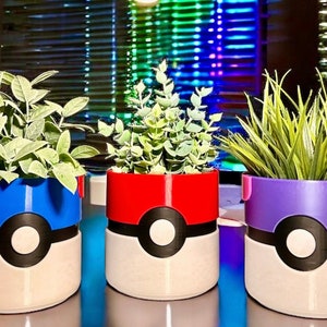 Puede incluir: Tres plantas en macetas de cerámica de colores con forma de Poké Ball. Las macetas son rojas, azules y moradas con una base blanca y detalles negros. Las plantas son verdes y frondosas.