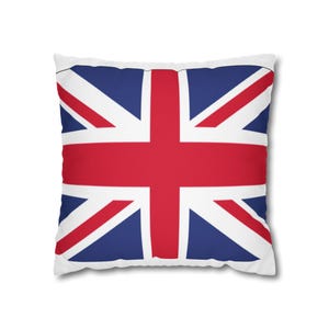 Pode incluir: Uma almofada quadrada com o design da bandeira Union Jack. O padrão consiste numa cruz vermelha ousada e faixas diagonais centradas num fundo branco, com secções triangulares azul-escuras a preencher os espaços restantes da almofada.