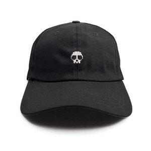May include: Black baseball cap with a pixelated white skull embroidered on the front. The cap has a curved brim and a fabric strap closure on the back. The cap is made of a durable, woven fabric.