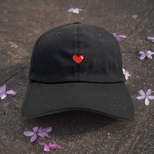 May include: Black baseball cap with a red pixelated heart embroidered on the front. The cap has a curved brim and a classic six-panel design. A stylish accessory for casual wear.