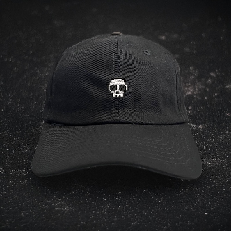 May include: Black baseball cap with a white pixelated skull design embroidered on the front. The cap has a curved brim and an adjustable strap at the back. The hat is made of a durable fabric.