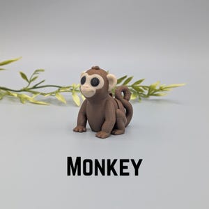 May include: A brown monkey figurine with a light-colored face and large black eyes. The word "MONKEY" is printed in bold black letters below the figurine. The monkey is sitting on a light gray surface with green and yellow leaves in the background.
