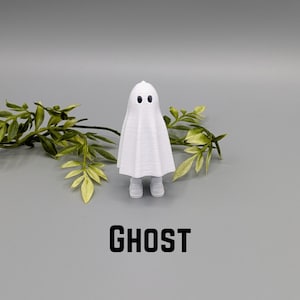 May include: A white ghost figurine with two black eyes and a draped sheet design. The word "GHOST" is printed in bold black letters at the bottom. The figurine is standing on a gray surface with green leafy sprigs in the background.