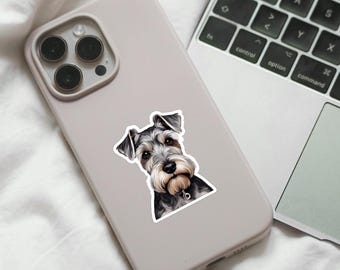 Peut inclure: Autocollant représentant un Schnauzer. L'illustration détaillée montre un chien avec un pelage gris et noir, une barbe et des sourcils distinctifs. L'autocollant est placé sur une coque de téléphone grise claire, avec un clavier d'ordinateur portable en arrière-plan.