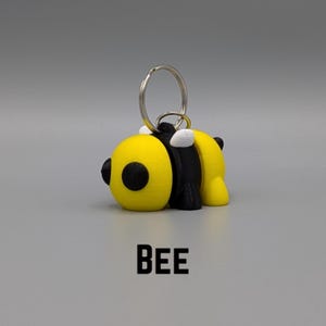 May include: A yellow and black bee keychain with white accents. The bee is in a 3D style and has a silver key ring.