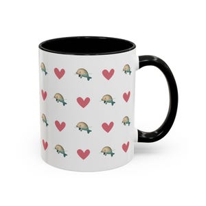 May include: White ceramic mug with a black interior and handle. The mug features a repeating pattern of red hearts and cartoon narwhals. The narwhals are light blue with a tan horn and belly. A fun and whimsical design.
