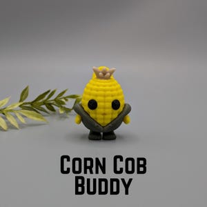 May include: A yellow corn cob figurine with black eyes, a crown, and green husk. The text "CORN COB BUDDY" is displayed in black block letters at the bottom of the image. The figurine is set against a gray background.