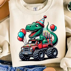 Może przedstawiać: Biała bluza z zielonym dinozaurem w czapce urodzinowej, jadącym czerwonym monster truckiem z kolorowymi balonami i świeczkami. Dinozaur uśmiecha się i ma radosny wyraz twarzy.