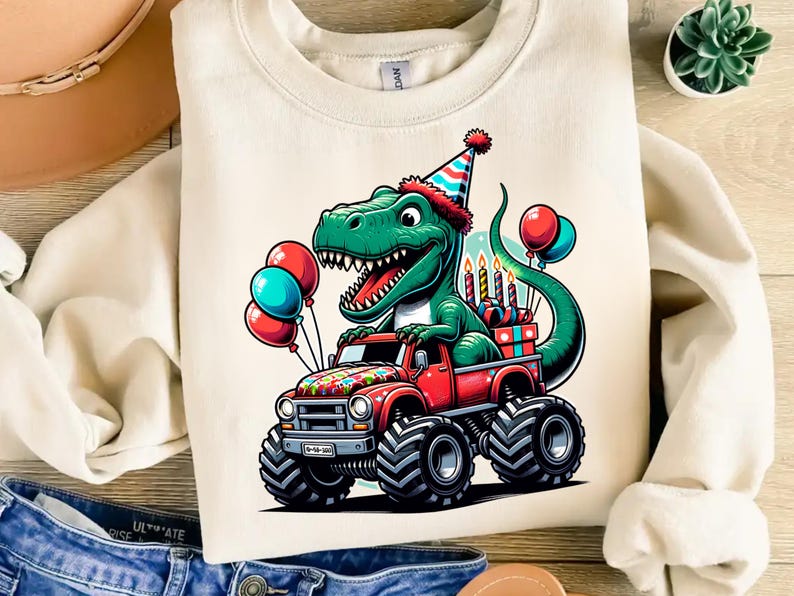 Może przedstawiać: Biała bluza z zielonym dinozaurem w czapce urodzinowej, jadącym czerwonym monster truckiem z kolorowymi balonami i świeczkami. Dinozaur uśmiecha się i ma radosny wyraz twarzy.