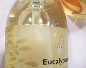 May include: Clear glass bottle with a white pump top and a yellow label. The label has a green leafy design and the text "TRINKETS Eucalyptus Net Wt. (100ml)"