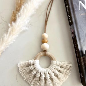 May include: A macrame wall hanging featuring a circular wooden ring adorned with off-white cotton cord knotted into a fan-shaped fringe. Above the ring, a brown leather cord holds two smooth wooden beads and one textured tan bead.