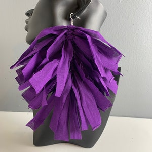 May include: Purple fabric tassel earrings. The earrings are long and dangle from the earlobe. The tassels are made of many small pieces of fabric that are tied together.