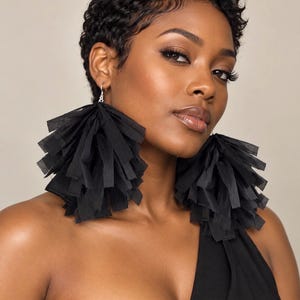 May include: A pair of large, black, fringe-style statement earrings. Each earring consists of numerous thin, layered strips of black fabric attached to a silver-toned metal hook, creating a voluminous, textured, and dangling tassel appearance.