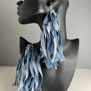 May include: A pair of denim fringe earrings. The earrings are made of strips of light and medium blue denim fabric, creating a layered, textured effect. The earrings hang from silver-colored fish hook ear wires. The earrings are displayed on a black mannequin head.