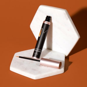 May include: A black and rose gold lash and brow growth serum bottle with a black applicator brush. The bottle is on a white marble hexagonal platform.