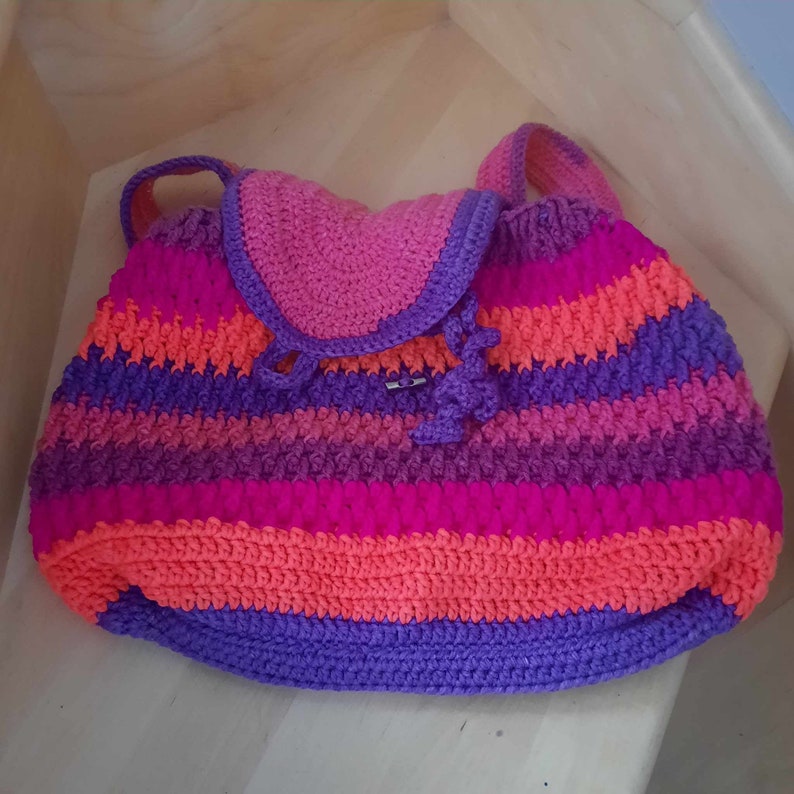 May include: A colorful crocheted backpack with a pink, orange, purple, and blue striped pattern. The backpack has a flap with a small buckle.