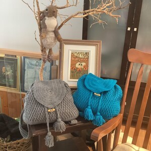 May include: Two crocheted backpacks with wooden toggles and tassels. One backpack is gray and the other is turquoise blue. The backpacks are sitting on a wooden table with a stuffed animal in the background.