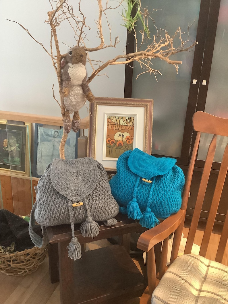 May include: Two crocheted backpacks with wooden toggles and tassels. One backpack is gray and the other is turquoise blue. The backpacks are sitting on a wooden table with a stuffed animal in the background.