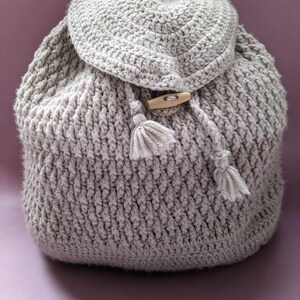 May include: A gray crocheted backpack with a wooden button and tassels. The backpack is made with a textured stitch pattern.
