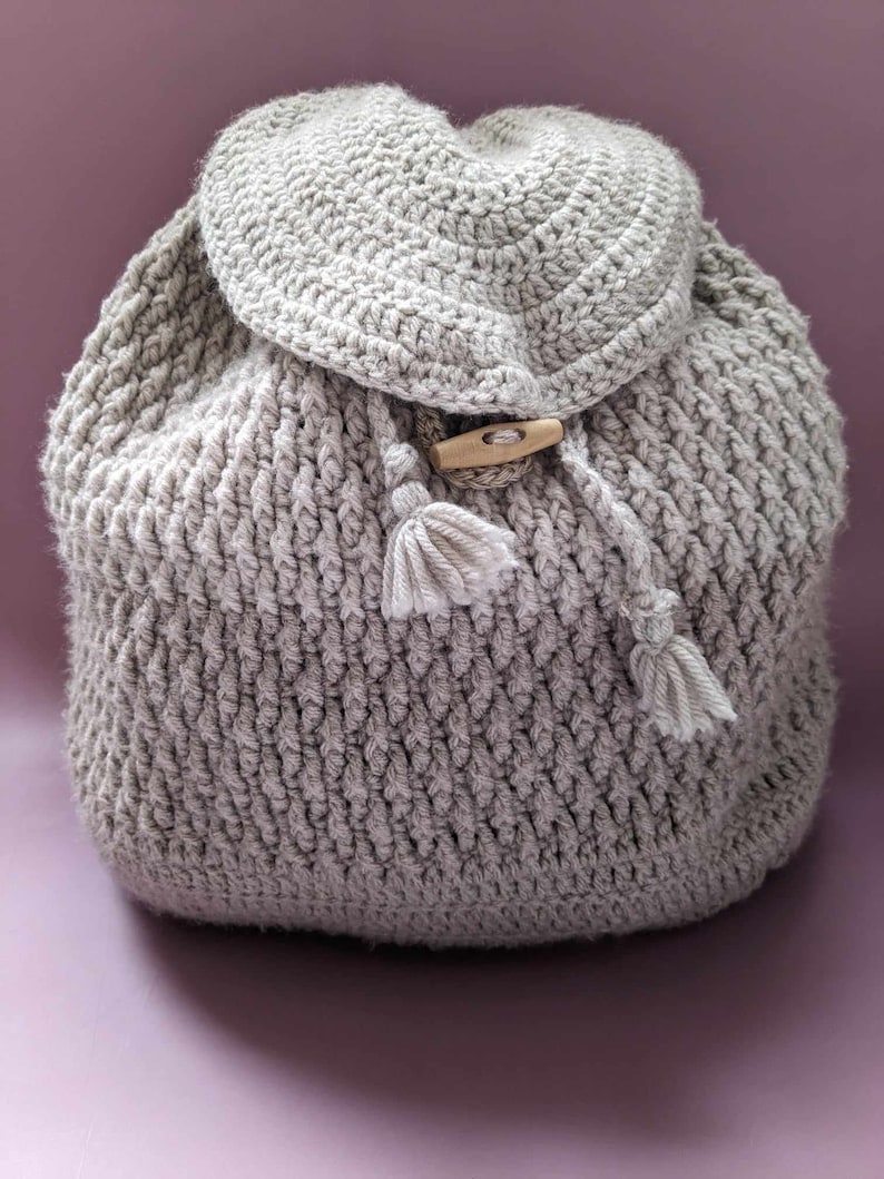 May include: A gray crocheted backpack with a wooden button and tassels. The backpack is made with a textured stitch pattern.