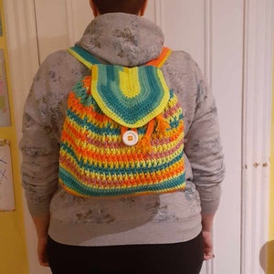 May include: A crocheted backpack with a colorful striped pattern in shades of green, yellow, orange, and pink. The backpack has a flap with a single white button closure and a tassel.