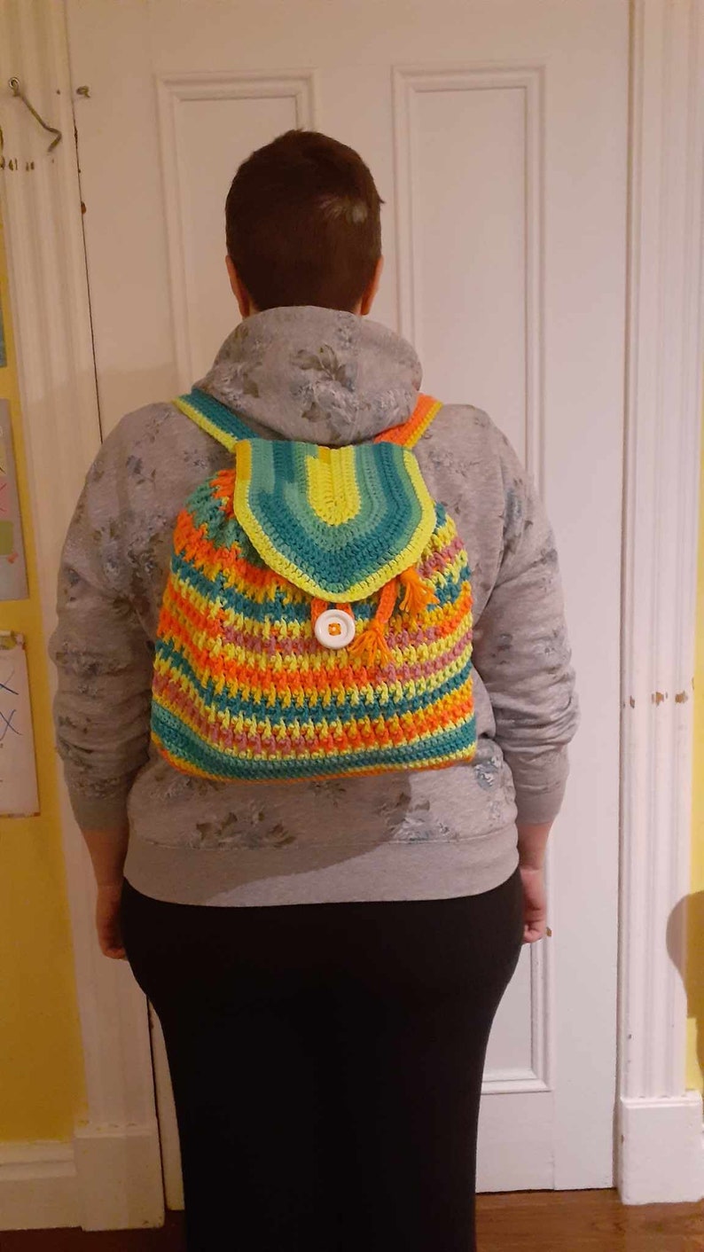 May include: A crocheted backpack with a colorful striped pattern in shades of green, yellow, orange, and pink. The backpack has a flap with a single white button closure and a tassel.