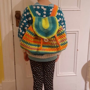 May include: A child wearing a white and blue polka dot sweater with a colorful crocheted backpack. The backpack has a bright green, yellow, orange, and pink design with a large button.