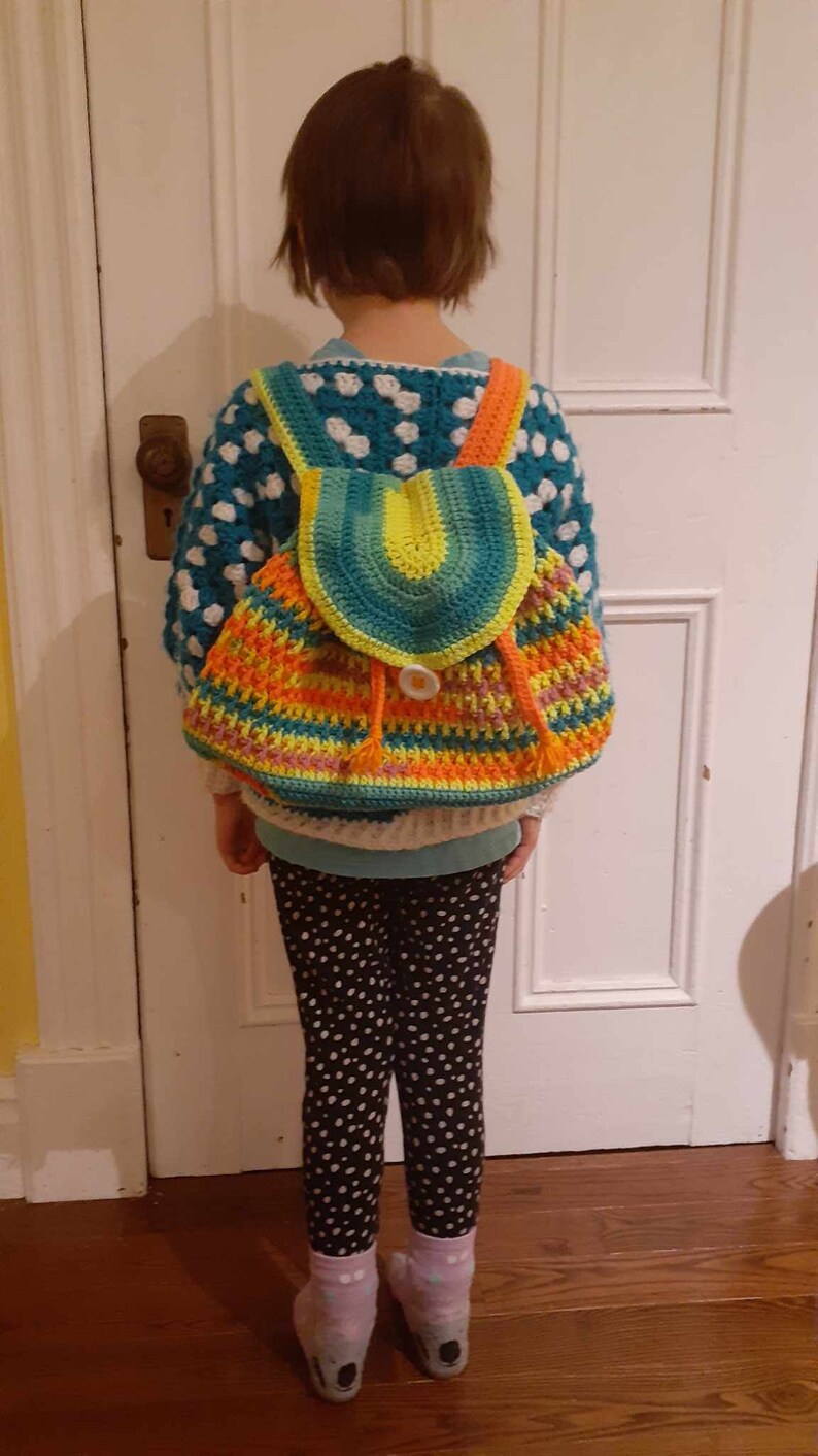 May include: A child wearing a white and blue polka dot sweater with a colorful crocheted backpack. The backpack has a bright green, yellow, orange, and pink design with a large button.