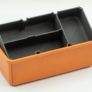 Puede incluir: Una caja de almacenamiento de pl&aacute;stico naranja con un inserto de pl&aacute;stico negro que tiene tres compartimentos. La caja est&aacute; vac&iacute;a.