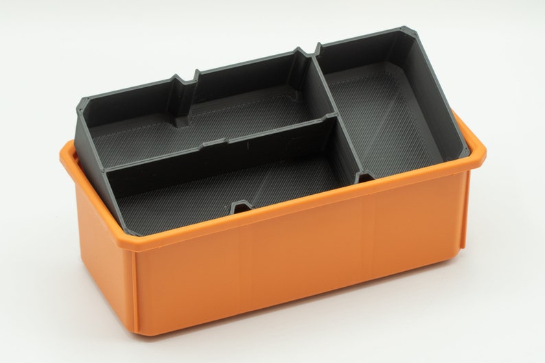 Puede incluir: Una caja de almacenamiento de pl&aacute;stico naranja con un inserto de pl&aacute;stico negro que tiene tres compartimentos. La caja est&aacute; vac&iacute;a.