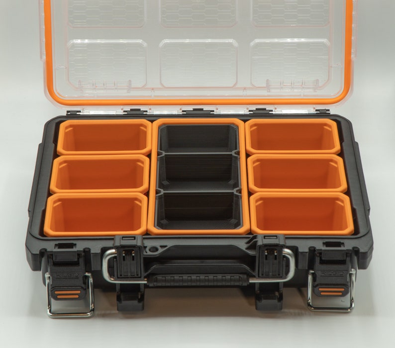 Puede incluir: Una caja de herramientas de pl&aacute;stico negra con un interior naranja. La caja de herramientas tiene varios compartimentos para organizar art&iacute;culos peque&ntilde;os. La tapa est&aacute; abierta, revelando los compartimentos vac&iacute;os.