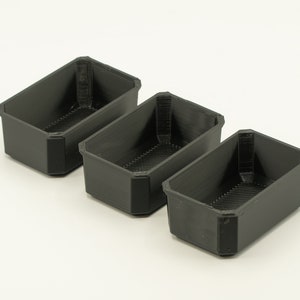 May include: Three black plastic rectangular containers with a textured surface. The containers are empty and are arranged in a row from left to right.
