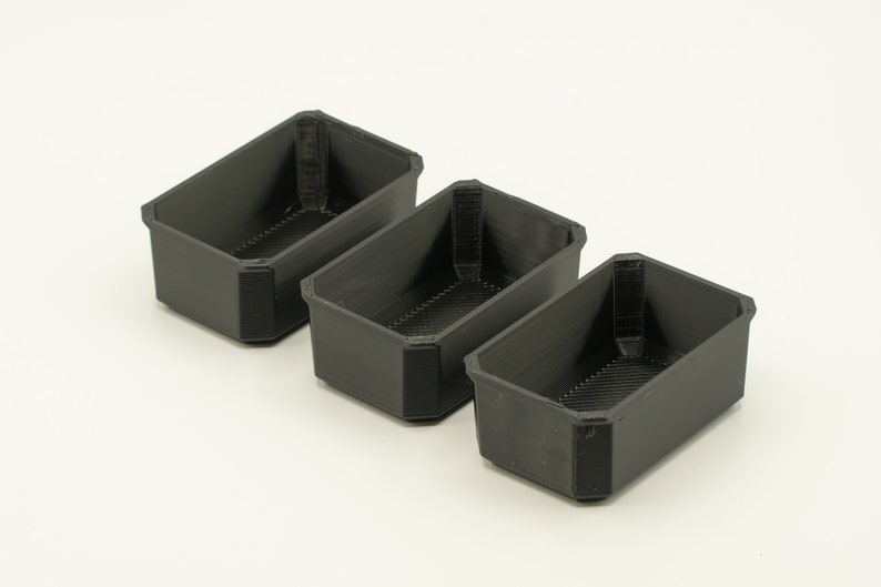 May include: Three black plastic rectangular containers with a textured surface. The containers are empty and are arranged in a row from left to right.