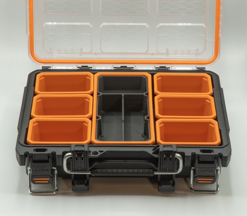 Puede incluir: Una caja de herramientas de pl&aacute;stico negra con un borde naranja y varios compartimentos extra&iacute;bles. La caja de herramientas est&aacute; abierta y los compartimentos est&aacute;n vac&iacute;os.