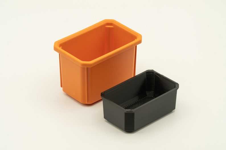 May include: Two plastic storage containers, one orange and one black. The orange container is larger and is sitting on a white surface. The black container is smaller and is sitting inside the orange container.