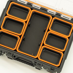 Puede incluir: Una caja de almacenamiento de pl&aacute;stico negro con un borde naranja y m&uacute;ltiples compartimentos. La caja est&aacute; abierta y los compartimentos est&aacute;n vac&iacute;os.