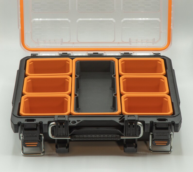 Puede incluir: Caja de herramientas de pl&aacute;stico negra y naranja con tapa transparente y varios compartimentos extra&iacute;bles. La caja de herramientas est&aacute; abierta y los compartimentos est&aacute;n vac&iacute;os.