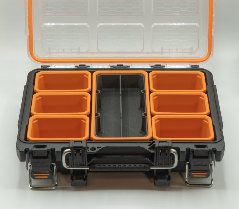 Puede incluir: Una caja de herramientas de pl&aacute;stico negra con varios compartimentos naranjas. La caja de herramientas tiene una tapa transparente y una manija de transporte.