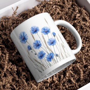 May include: White ceramic mug with a watercolour design of blue cornflowers and wheat. The mug has a white handle and is in a white box with brown paper. A floral-themed gift, ideal for tea or coffee.