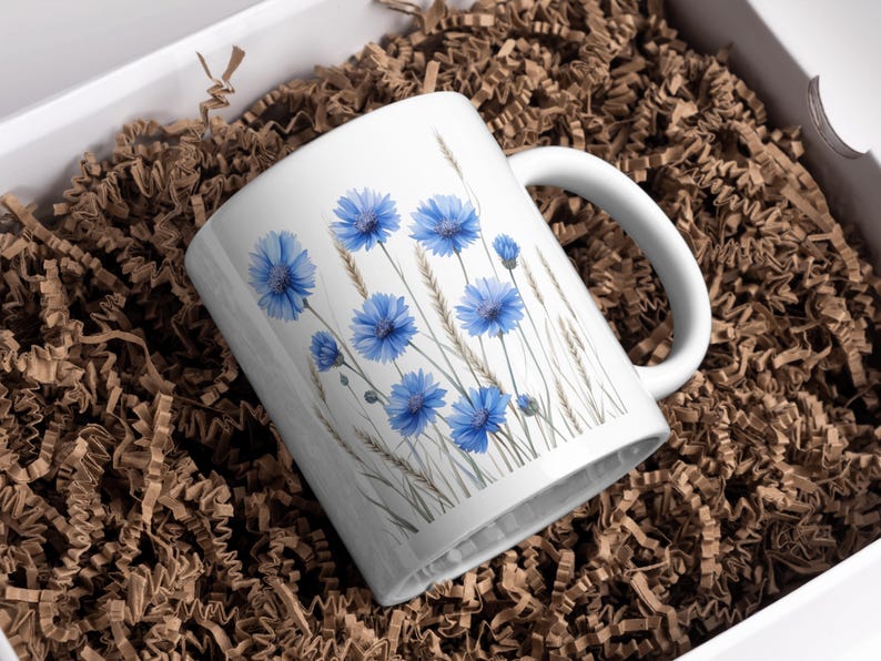 May include: White ceramic mug with a watercolour design of blue cornflowers and wheat. The mug has a white handle and is in a white box with brown paper. A floral-themed gift, ideal for tea or coffee.