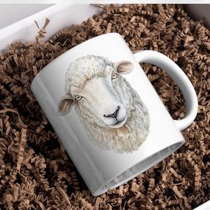 May include: White ceramic mug featuring a watercolor illustration of a sheep's head. The sheep has fluffy white wool, brown ears, and a black nose. The mug is presented in a white box with brown paper filler.